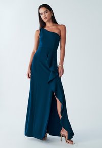 Teal One-Shoulder-Kleid mit tailliertem Oberteil, Rüschendetail und einem Oberschenkel hohen Schlitz. Aus glattem Stoff gefertigt. Model trägt goldene Accessoires.
