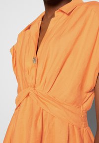 Gros plan sur une personne portant une robe orange avec un col, un seul bouton visible et un détail en tissu torsadé à la taille.