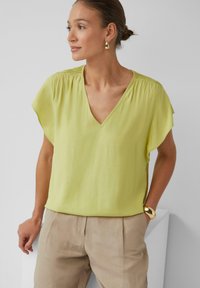 Lichtgele blouse met een V-hals, korte mouwen en geplooide schouderdetails. Gaan gepaard met beige broek en gouden accessoires.