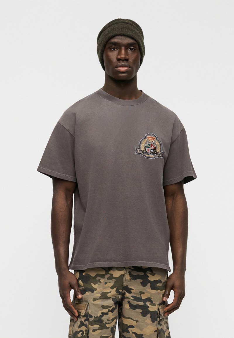 Γκρι βαμβακερό T-shirt με χαλαρή εφαρμογή, με κεντημ ένο λογότυπο στο αριστερό στήθος. Συνδυασμένο με παντελόνια καμουφλάζ cargo και πλεκτό σκούφο.