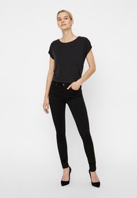 Top preto de manga curta combinado com jeans skinny pretos e sapatos de salto agudo pretos. O conjunto apresenta um corte descontraído e uma textura de tecido suave.