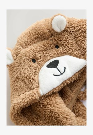 Capuche en peluche brun avec des yeux noirs brodés, un nez, et une bouche souriante sur une douce fourrure blanche, faisant partie d'un vêtement ou d'une couverture en peluche.