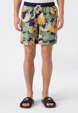 Hombre con pantalones cortos multicolor con estampado abstracto de camuflaje y cintura con cordón, y sandalias negras tipo slide con logotipo, de pie sobre un fondo liso.