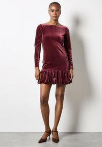 Bordowa sukienka z weluru z długimi rękawami i falbaną u dołu. Zwężający się krój, gładka tekstura i dekolt w łódkę. Elegancki i młodzieńczy wygląd.