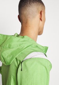 Veste légère verte zippée avec capuche. Caractérisée par un tissu texturé et une poche cachée avec une doublure blanche à petits pois.