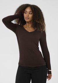 Brun langærmet top med V-halsdesign, lavet af blødt stof. Har tætsiddende ærmer og en glat tekstur. Kombineret med sorte bukser.