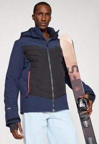 Marineblauer Skijacke mit schwarzem gepolstertem Vorderteil, Reißverschluss und roten Akzenten. Das Modell hält einen Ski mit strukturierten Oberflächen und bedruckten Grafiken.