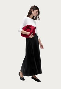 Black pleated skirt, white long-sleeve top, carrying a textured red handbag. Black flats complete the outfit, emphasizing contrast in colors.