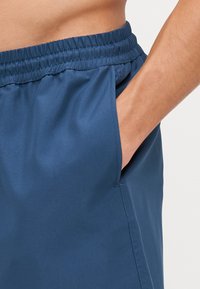Shorts mit elastischem Bund aus dunkelblauem Stoff, ausgestattet mit Seitentaschen und einer glatten Textur, die für Komfort und Bewegungsfreiheit sorgen.