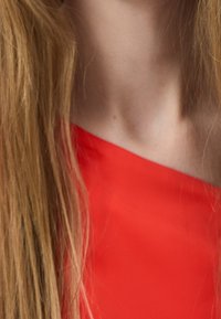 Indumento rosso, scollato e senza spalle, con una texture liscia. Capelli biondi lunghi e lisci parzialmente visibili accanto a una pelle chiara. Nessun motivo evidente.