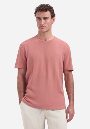 Man with curly hair wearing a textured dusty pink short-sleeve shirt and light beige pants, standing with hands in pockets.