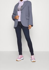 Blazer gris con mangas anchas, dos bolsillos frontales, superpuesto sobre una sudadera de color púrpura claro. Jeans oscuros ajustados y zapatillas rosas con suelas blancas.