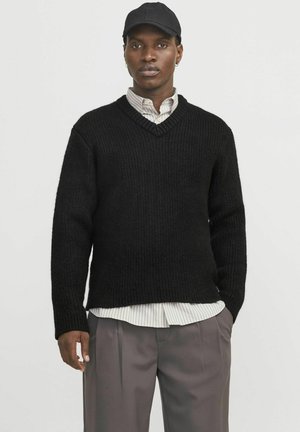 Maglione nero a maglia con scollo a V e texture a coste, sovrapposto a una camicia bianca a righe. I pantaloni grigi completano l'outfit.
