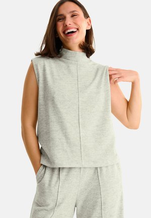 Femme souriante, portant un top sans manches gris clair à col montant et un short assorti, les mains dans les poches, sur fond blanc.