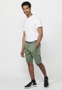 Grüne Cargo-Shorts mit Seitentaschen und umgeschlagenen Säumen, kombiniert mit einem schlichten weißen T-Shirt und schwarzen Sneakers. Aus glattem Baumwollmaterial.
