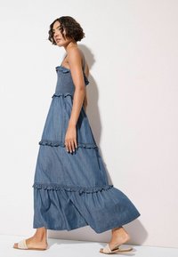Denim maksi obleka z večplastnimi volančki in nabranim bodicem. Svetlo modra barva, tekoča tkanina in nastavljive naramnice.
