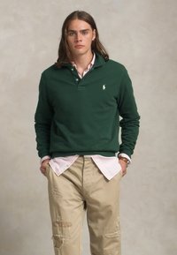 Pull vert en coton avec un col et un petit logo, superposé sur une chemise à rayures roses. Pantalon beige avec des détails usés.