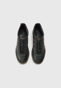 Melni ādas sporta apavi ar brūnām svītrām, teksturētu virsmu, šņorējamu dizainu, gumijas zoli un zelta logotipa detaļu uz mēles.