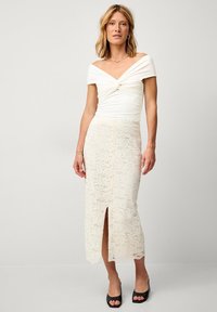 Haut blanc décolleté avec détail torsadé, associé à une jupe en dentelle crème midi avec des motifs fleuris et une fente sur le devant. Escarpins noirs.