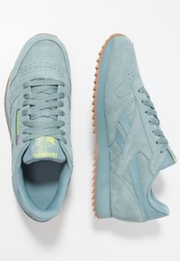 Ljusblå mockaskor med texturerade accenter, gummisulor i karamellfärg och kontrasterande detaljer, inklusive en logotyp och sömmönster.