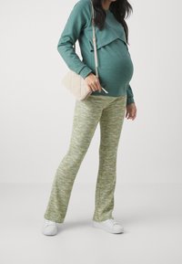 Grön sweatshirt med ett lager på framsidan, i kombination med mönstrade utsvängda byxor i grönt och vitt. Vita sneakers och en beige axelväska.