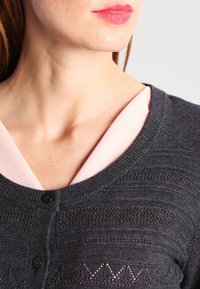 Mörkgrå stickad cardigan med ett texturerat mönster, rund halsringning och svart knäppning, lager på lager över en ljusrosa topp.