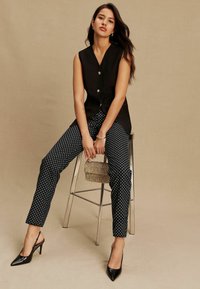 Must varrukateta pluus ees nupu ja must-valge täpilise mustriga püksid. Modell istub pingil, jalas mustad terava ninaga kontsakingad.