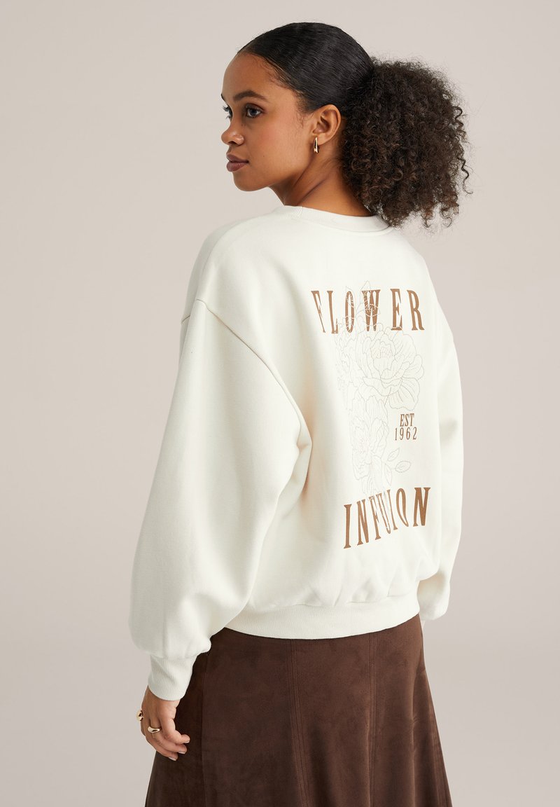 Junge Frau mit lockigem Haar in einem weißen, übergroßen Sweatshirt mit Blumenmuster und braunem Rock, leicht zur Seite gedreht.