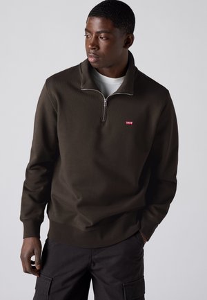 Jeune homme portant un sweat-shirt marron foncé à fermeture éclair quart, avec un logo rouge Levi's, pantalon noir, regardant sur le côté sur un fond uni.