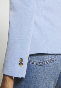 Manche de blazer bleu clair texturé avec deux boutons marron à côté de la poche arrière d'un jean denim bleu porté par une personne.