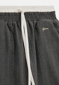 Pantaloni corti grigi a righe sottili con vita elastica, cordino bianco e un piccolo logo dorato "Guess" sul davanti. Dettagli in tessuto testurizzato.