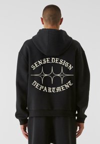 Fekete kapucnis pulóver, laza szabással, fehér „SENSE.DESIGN DEPARTMENT” felirattal és dekoratív vonalakkal a hátsó részen. Puha anyag, bordázott mandzsettákkal.