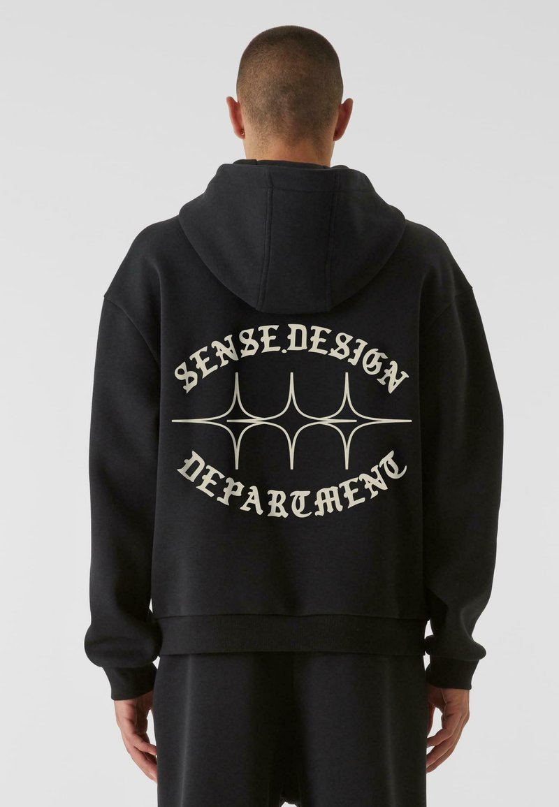 Fekete kapucnis pulóver, laza szabással, fehér „SENSE.DESIGN DEPARTMENT” felirattal és dekoratív vonalakkal a hátsó részen. Puha anyag, bordázott mandzsettákkal.
