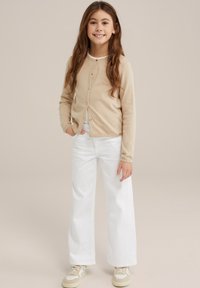 Bežs adīts kardigāns ar apaļu kaklu, priekšā pogām, kombinēts ar baltām platām biksēm un bežām kedām. Garas, viļņotas matu šķipsnas.