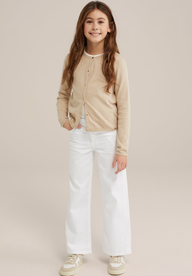 Beige pletený cardigan s kulatým výstřihem, předními knoflíky, v kombinaci s bílými širokými kalhotami a béžovými teniskami. Dlouhé, vlnité vlasy.