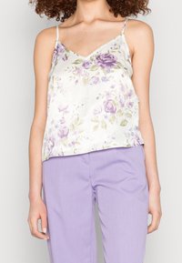 Femme portant un camisole en satin blanc à fleurs avec des roses violettes et un pantalon violet clair, se tenant devant un fond clair.