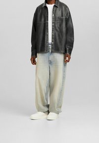 Černá košile z umělé kůže na knoflíky, světle modré volné džíny a bílé tenisky. Má kapsu na hrudi a kontrastní švy.