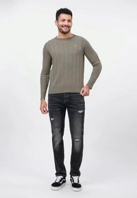 Hymyilevä mies, jolla on päällään oliivinvihreä ribbineule, kuluneet mustat farkut ja mustat lenkkarit, joissa on valkoiset nauhat. Hän seisoo valkoista taustaa vasten.