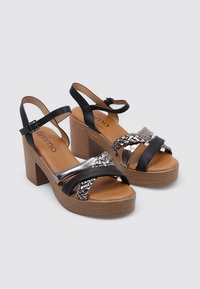 Sandalias de plataforma negras y plateadas con un detalle de cinta trenzada, que cuentan con un tacón de bloque de madera y una correa en el tobillo para mayor soporte.