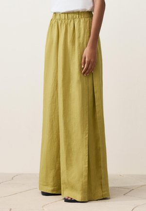 Oseba oblečena v široke, visokoločajne lanu rumene hlače z elastičnim pasom in črne sandale z odprtimi prsti, stoječa na svetlem tlakovcu.