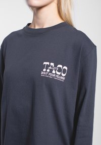 Langärmliges navy Blau T-Shirt aus Baumwolle, mit einem rosa grafischen Druck auf der linken Brust, der Text und Logos enthält. Glatte Textur, lockere Passform.