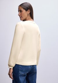 Helles beiges Langarmoberteil mit einem runden Ausschnitt und Puffärmeln, kombiniert mit blauen Jeans. Glatte Textur, minimalistisches Design.
