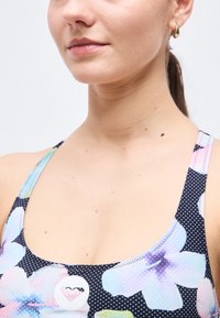 Femme portant un débardeur floral coloré à encolure arrondie et une petite boucle d'oreille en or, sur un fond blanc uni.