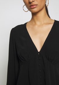 Robe noire avec un décolleté en V profond, fermeture à boutons et détails froncés au niveau de la poitrine. Confectionnée en tissu doux et léger ; manches longues.