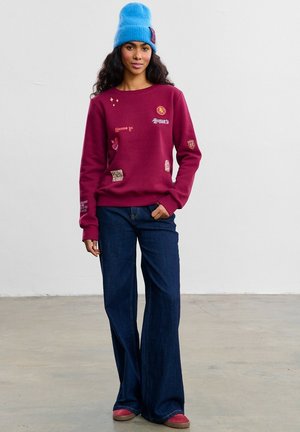 Bordowa bluza z kapturem z różnymi naszywkami, zestawiona z szerokimi, ciemnoniebieskimi dżinsami oraz jasnoniebieską dzianinową czapką. Obuwie w kolorze czerwonym.
