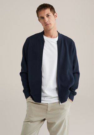 Man met kort bruin haar, gekleed in een marineblauwe zip-up jas, een wit T-shirt en beigekleurige broek, staande met handen in de zakken.