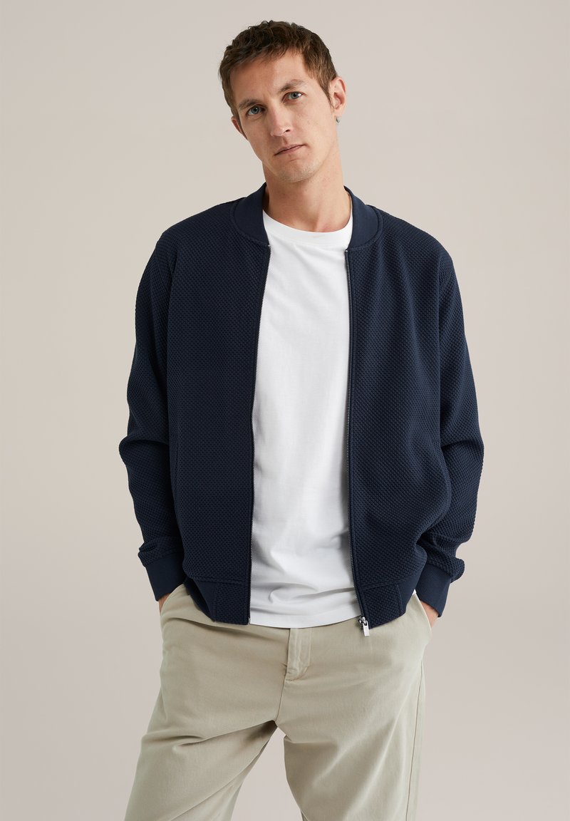 Man met kort bruin haar, gekleed in een marineblauwe zip-up jas, een wit T-shirt en beigekleurige broek, staande met handen in de zakken.