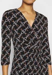 Robe portefeuille noire à manches trois-quarts, présentant un motif géométrique de cercles entrelacés en nuances de blanc et de brun. Tissu texturé.