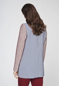 Long-sleeve top featuring navy and white geometric pattern, sheer burgundy sleeves, and a rounded neckline. Smooth texture and relaxed fit.