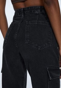 Sono mostrati pantaloni in denim nero visti da dietro, con grandi tasche e una vita alta. Il tessuto ha un aspetto leggermente sbiadito.